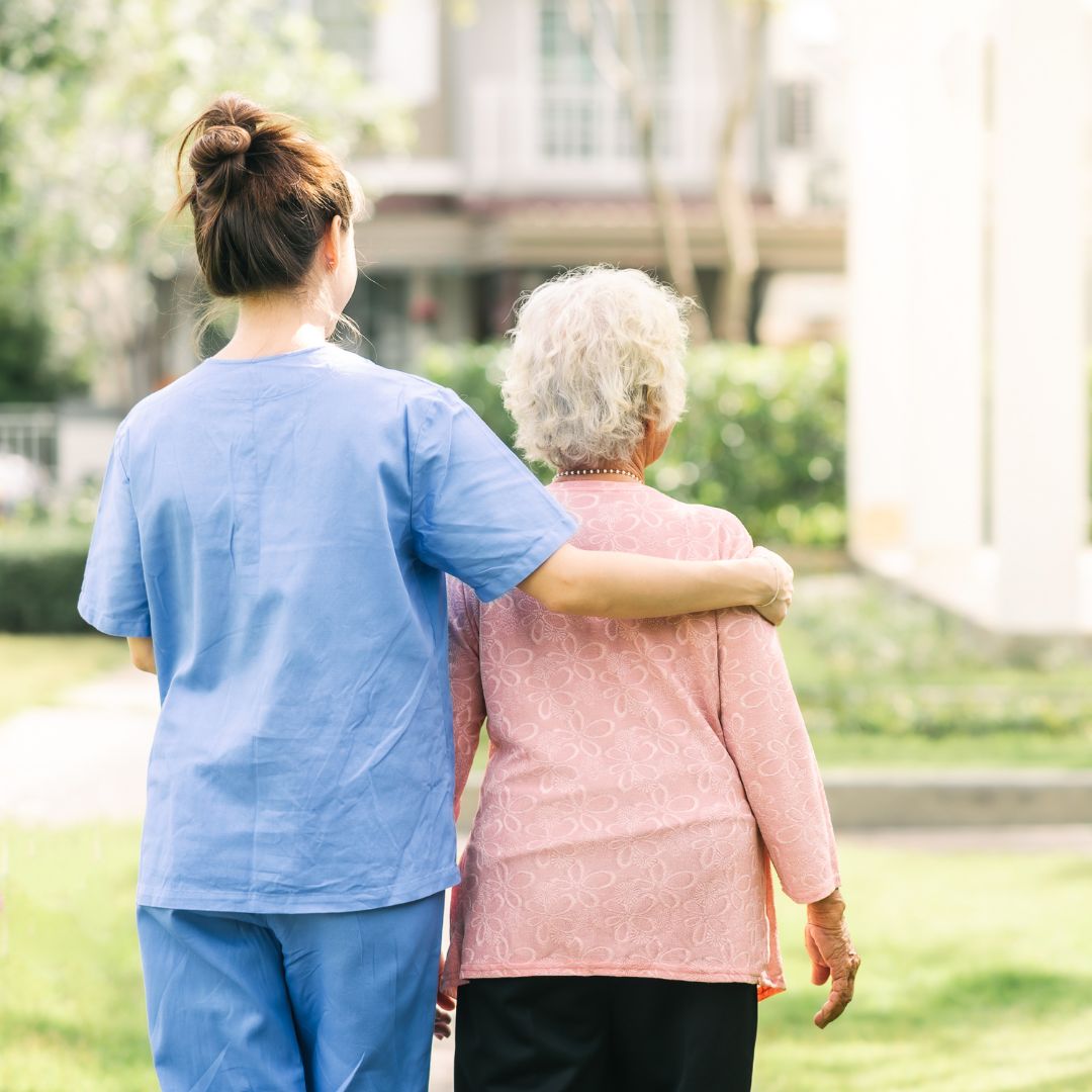 Nurse wearing blue scrubs walking with arm around elderly woman in peach shirt and black pants