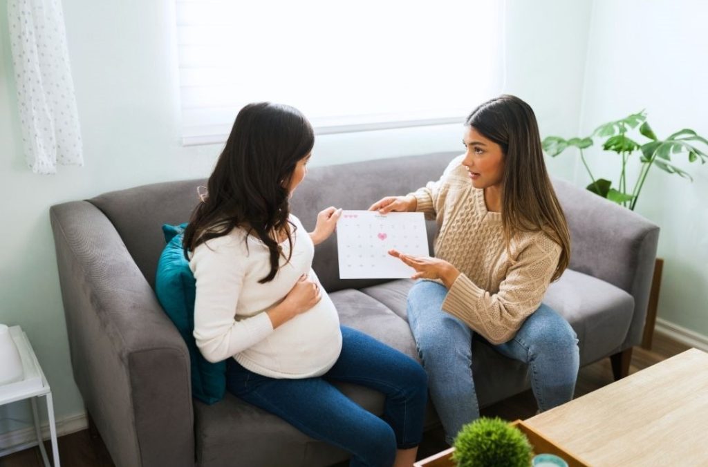 A pregnant woman sits on a couch talking with another woman who is holding a document and gesturing as she speaks.